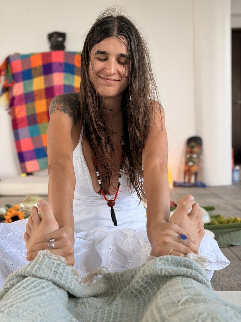 Johannie in white dress smiling, holding both feet in a seated stretch with mala beads at her chest