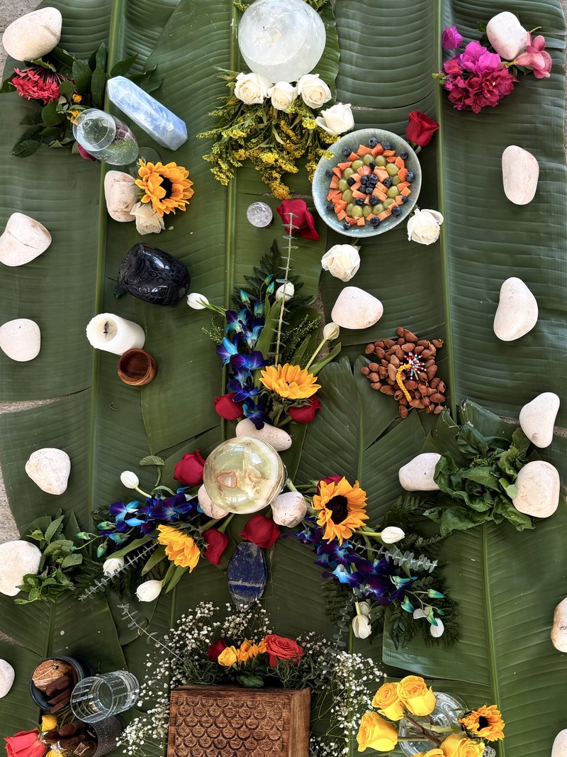 Altar overhead on banana leaves with sunflowers, blue orchids, red and white roses, crystals, fruit bowl, and stones